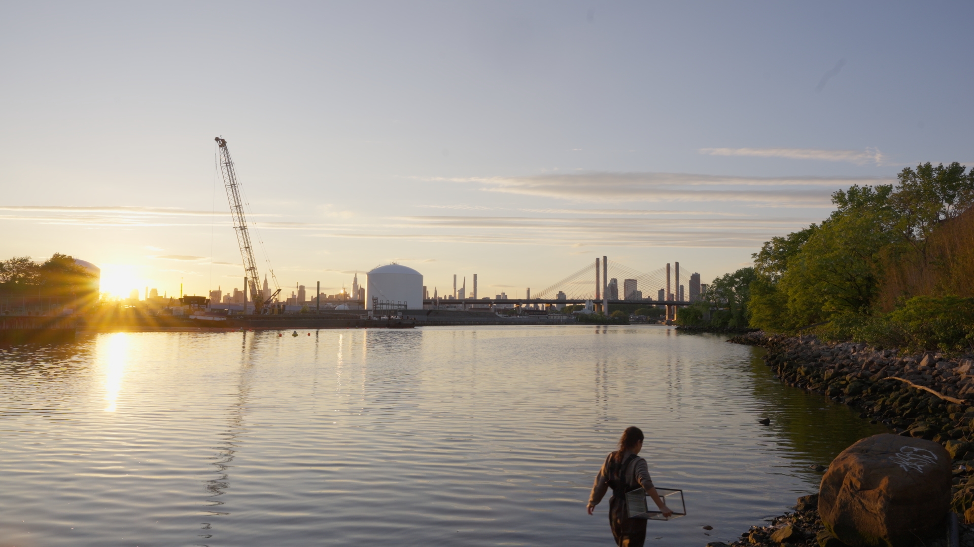 A figure wades through Newtown Creek at sunset with the Manhattan skyline behind