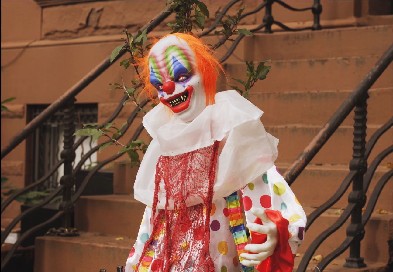 A grinning clown decoration with orange hair and a polka-dot costume, propped on a brownstone stoop