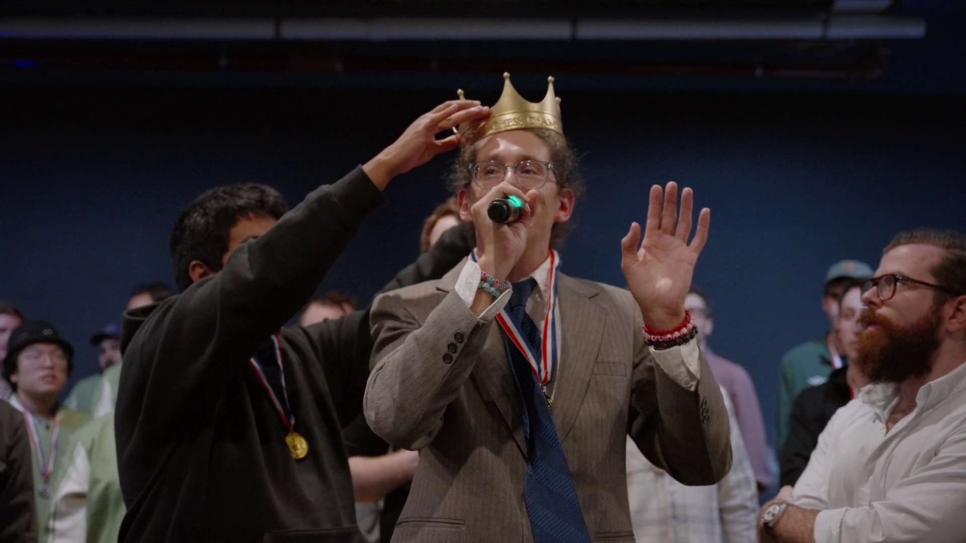 A newly crowned chess champion raises his hands as a gold paper crown is placed on his head