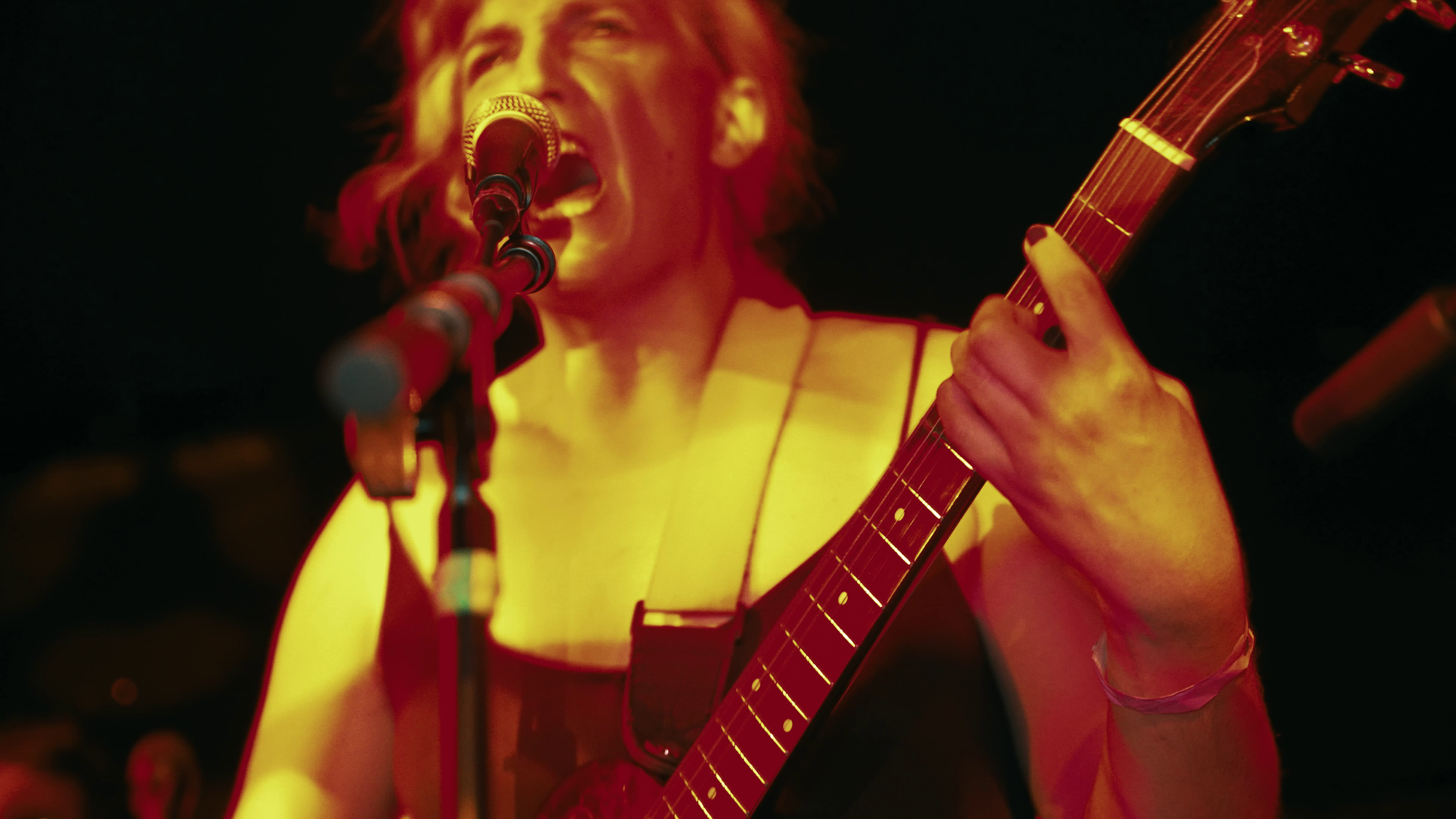 A guitarist bathed in red and yellow stage light, singing into a microphone