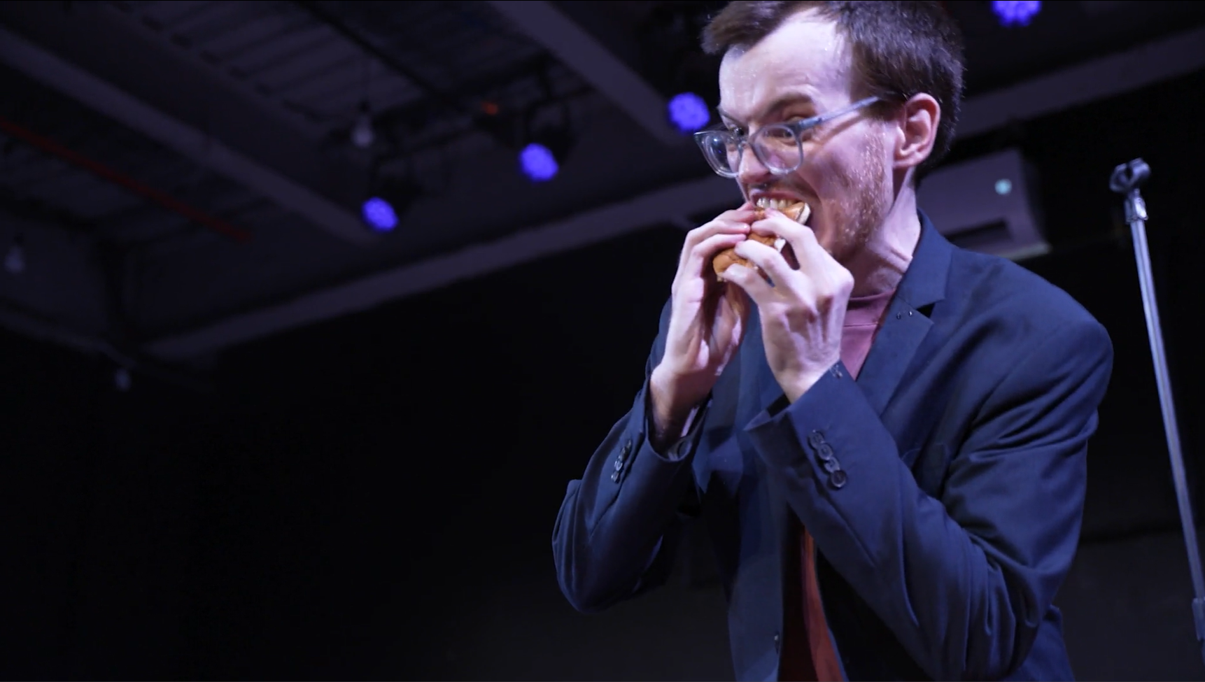 A contestant in a blazer biting into a hot dog under stage lights