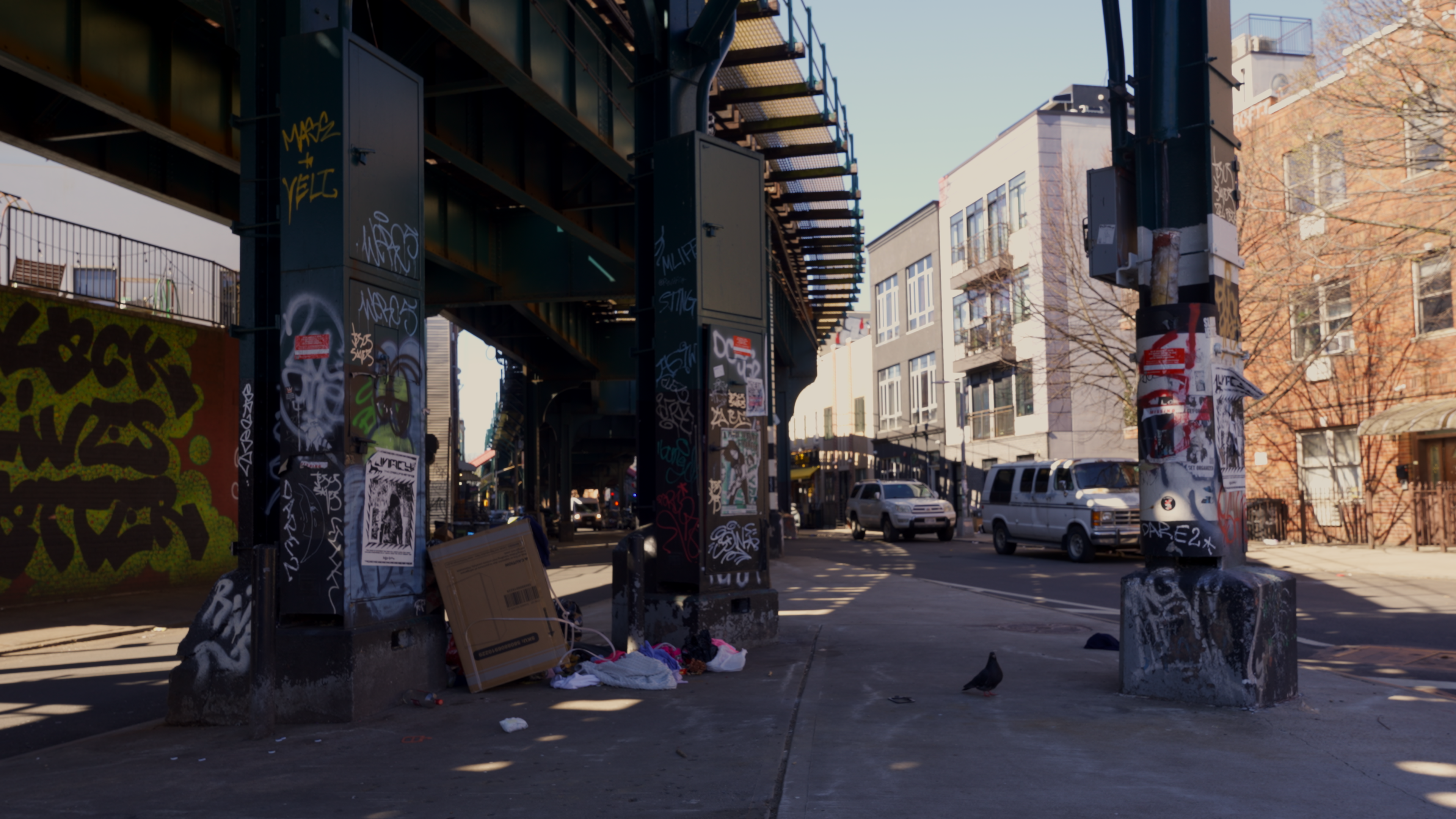 Graffiti-covered columns under the elevated train, a broken cabinet and bagged refuse left on the sidewalk