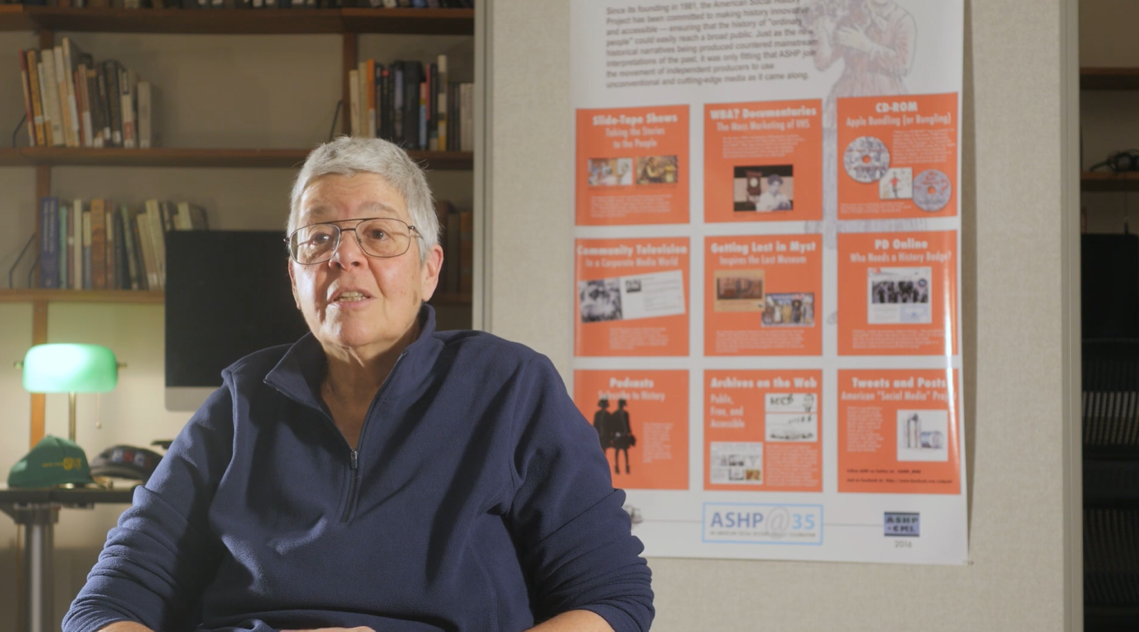 A historian speaking on camera in an office, seated in front of an orange ASHP at 35 timeline poster